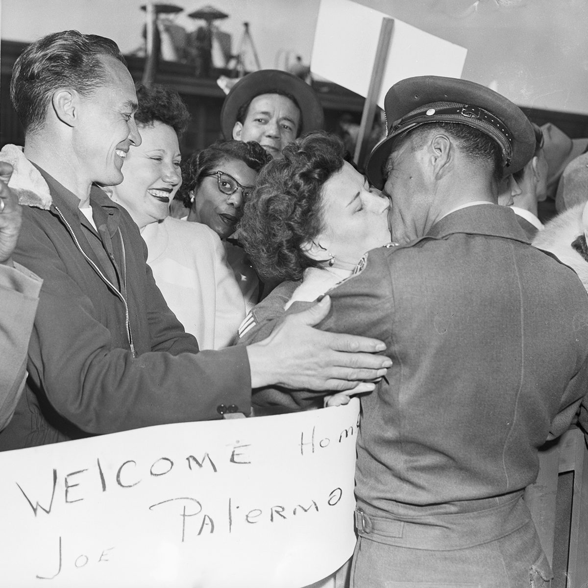 A uniformed soldier kisses a woman amid a joyful crowd. Smiling people surround them, and a hand-written sign reads "Welcome Home Joe Palermo." The scene conveys happiness and celebration.