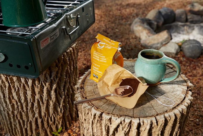 Coffee package and teal mug sit on a tree stump near a campfire pit; adjacent green camping stove visible. Text: "Coffee, sweet espresso."