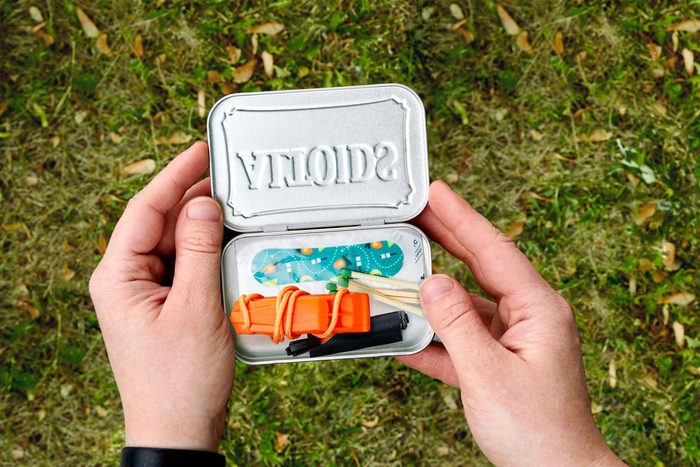 Hands hold an open mint tin containing various survival items, viewed against grassy ground.