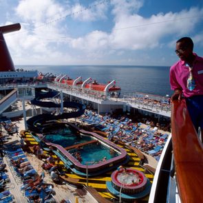 Bartender on the top deck of a Carnival Cruise ship holds two large colorful frozen drinks in hand, overlooking the pool area
