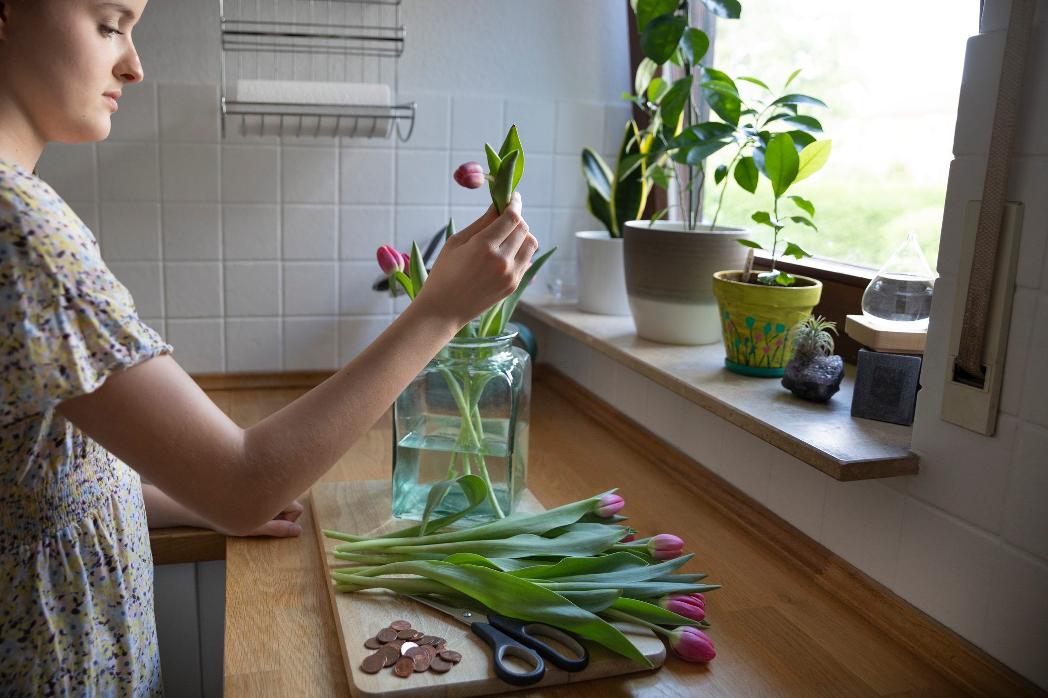 Woman arranging purple-pink tulips into a large glass jar part filled with water and with some copper coins ready to also add to the water