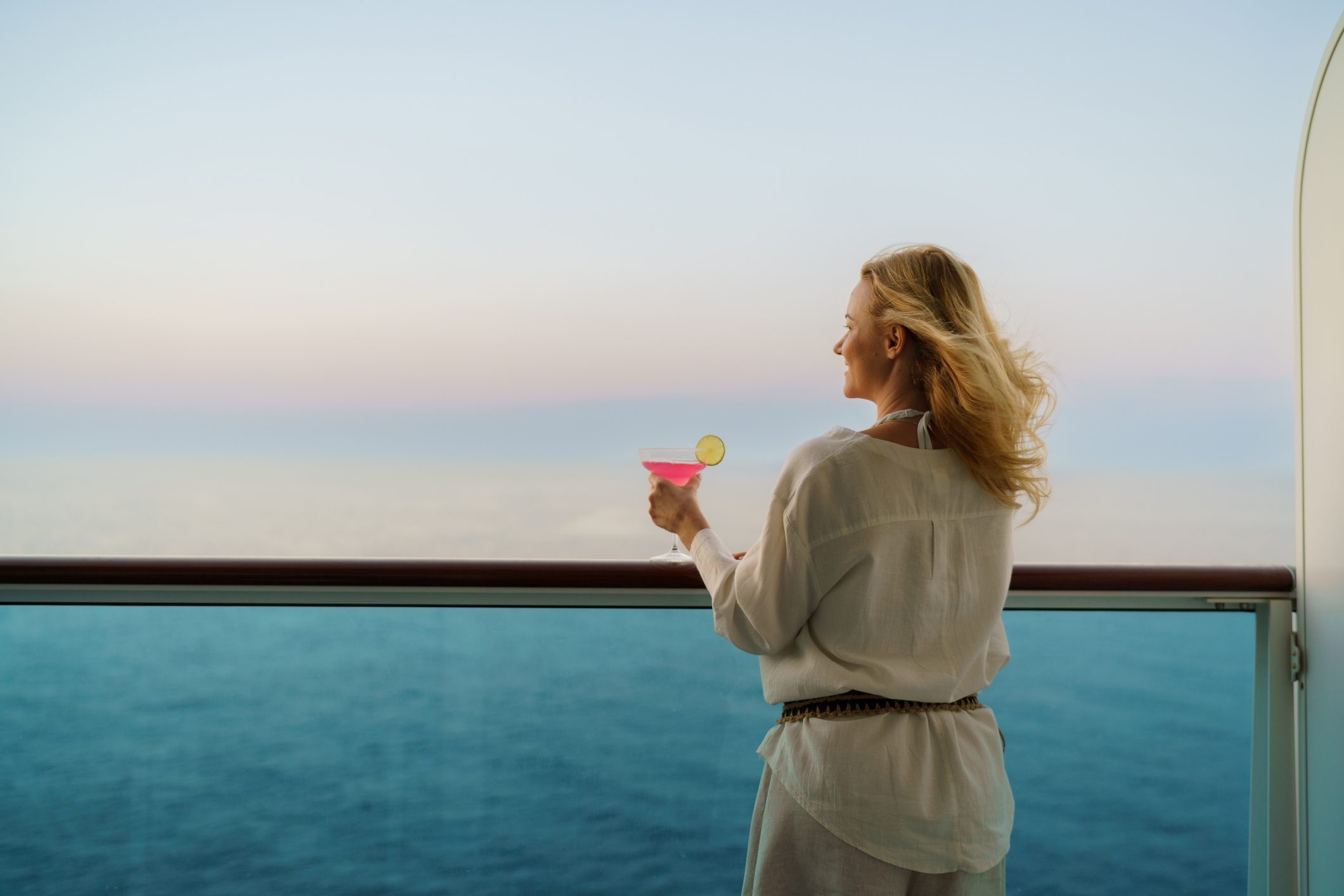 A woman with a cocktail in her hand is looking at the sea on the balcony of a cruise ship