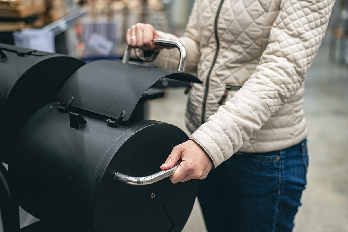 A person wearing a quilted beige jacket and jeans is opening the lid of a black metal smoker grill with both hands in an indoor setting.
